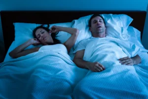 Ein schlafender Mann mit obstruktiver Schlafapnoe schnarcht laut im Bett, während seine gequälte Frau sich die Ohren zuhält